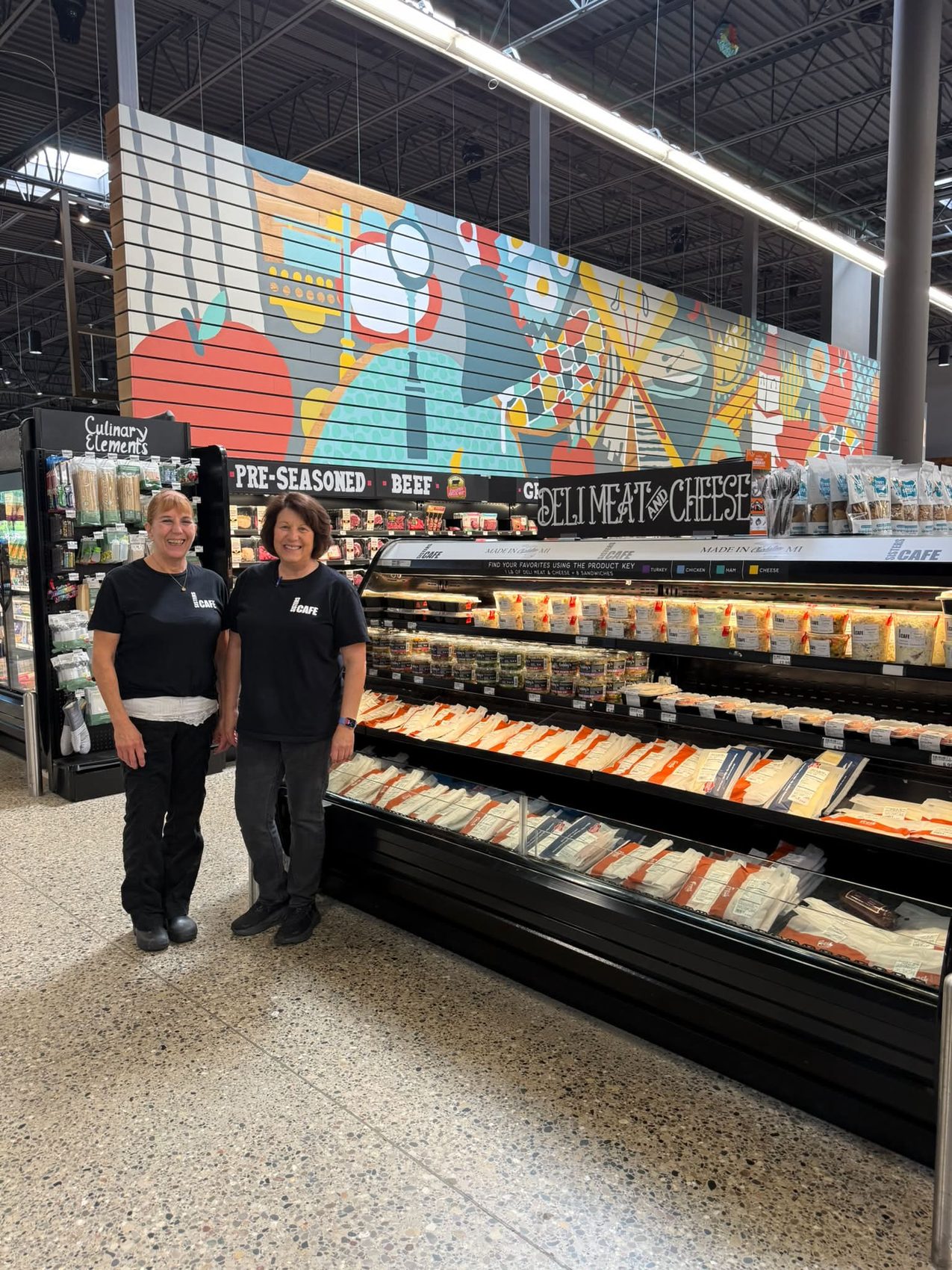 Two Sisters Cafe team members standing beside the stocked deli case at Meijer
