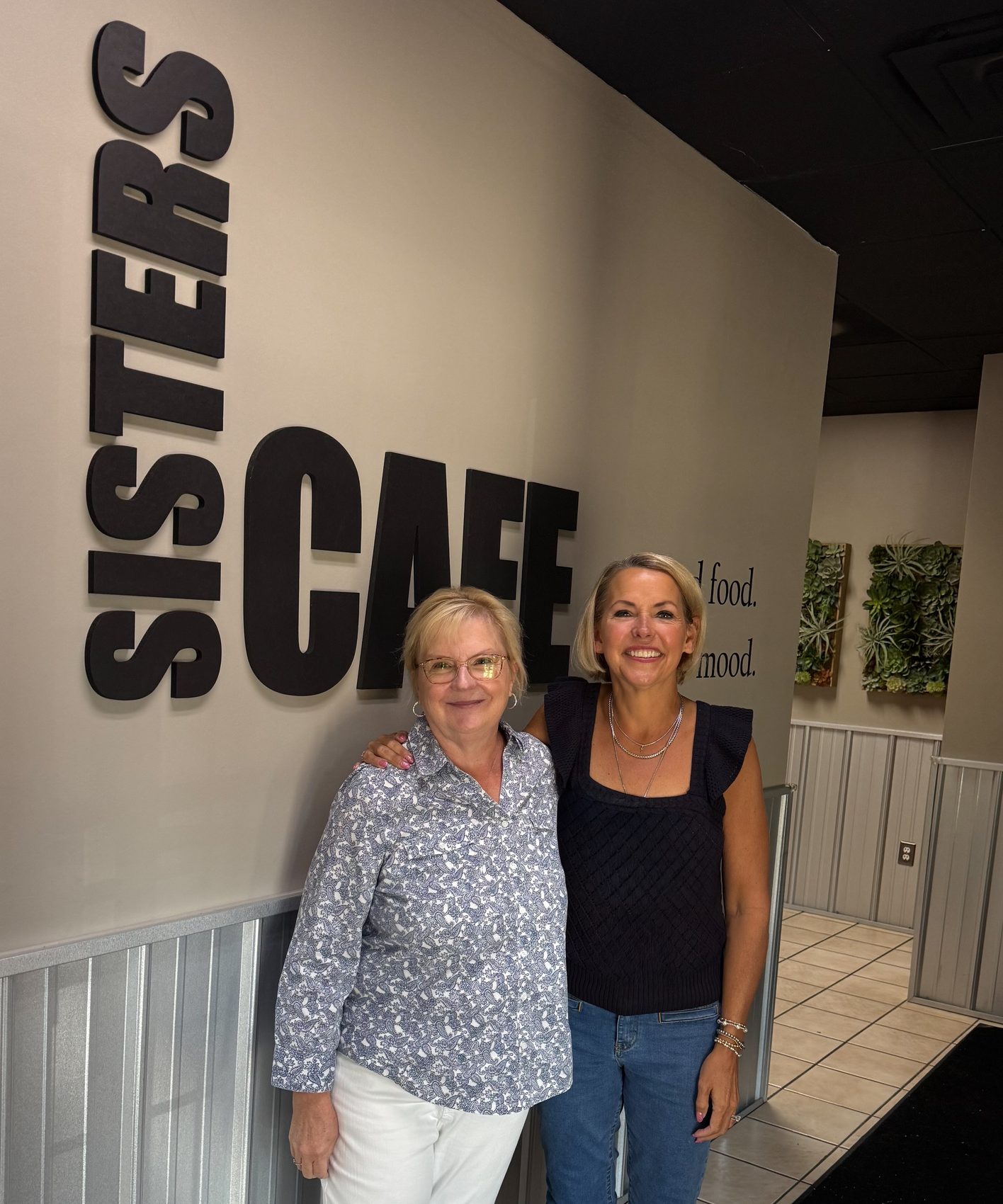 Two women standing together in front of the large black SISTERS CAFE lettering on the cafe wall